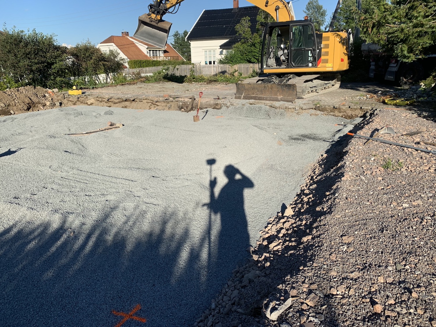 skygge av en person som gjoer innmaaling ses paa bakken hvor det er gjort grunnarbeid. Gravemaskin i andre enden.