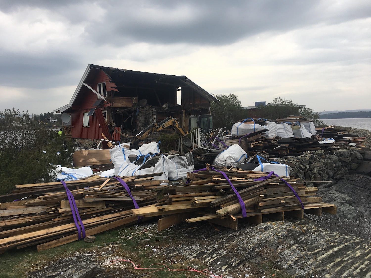 et halvrevet roedt hus foran vannet. Rivematerialer ligger i front.